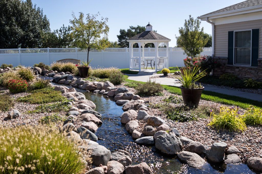 A beautiful garden with a stream flowing through rocks, surrounded by lush greenery and featuring a gazebo in the background.