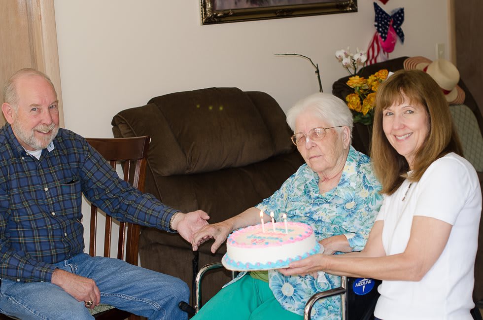 A family celebrates a birthday with an elderly woman holding a cake indoors.