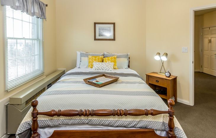 A cozy bedroom features a neatly made bed with neutral tones, a wooden nightstand, and a window providing natural light.
