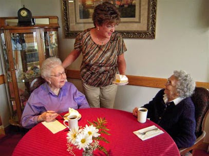 A caregiver serving cake to two elderly women during a social gathering at a cozy cafe.
