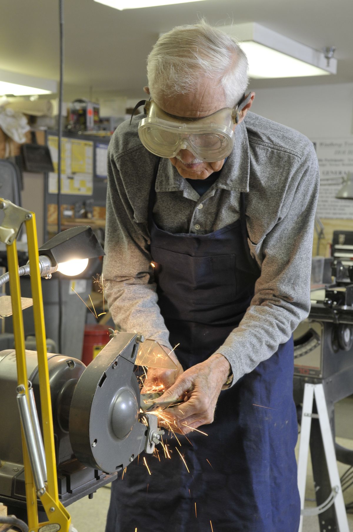 A person wearing safety glasses and an apron works with a grinding machine in a workshop.