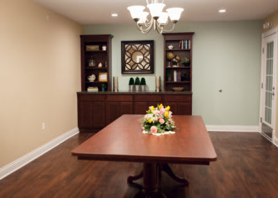 A dining room with a central wooden table, adorned with a floral centerpiece. Bookshelves and a decorative mirror enhance the background ambiance.