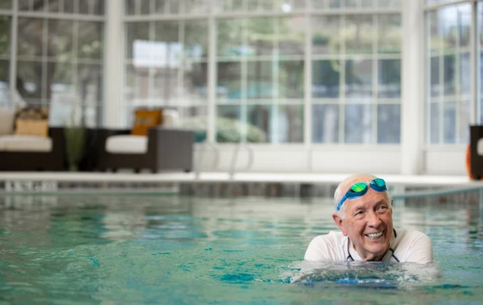 An elderly person enjoys swimming in an indoor pool, smiling with goggles on.