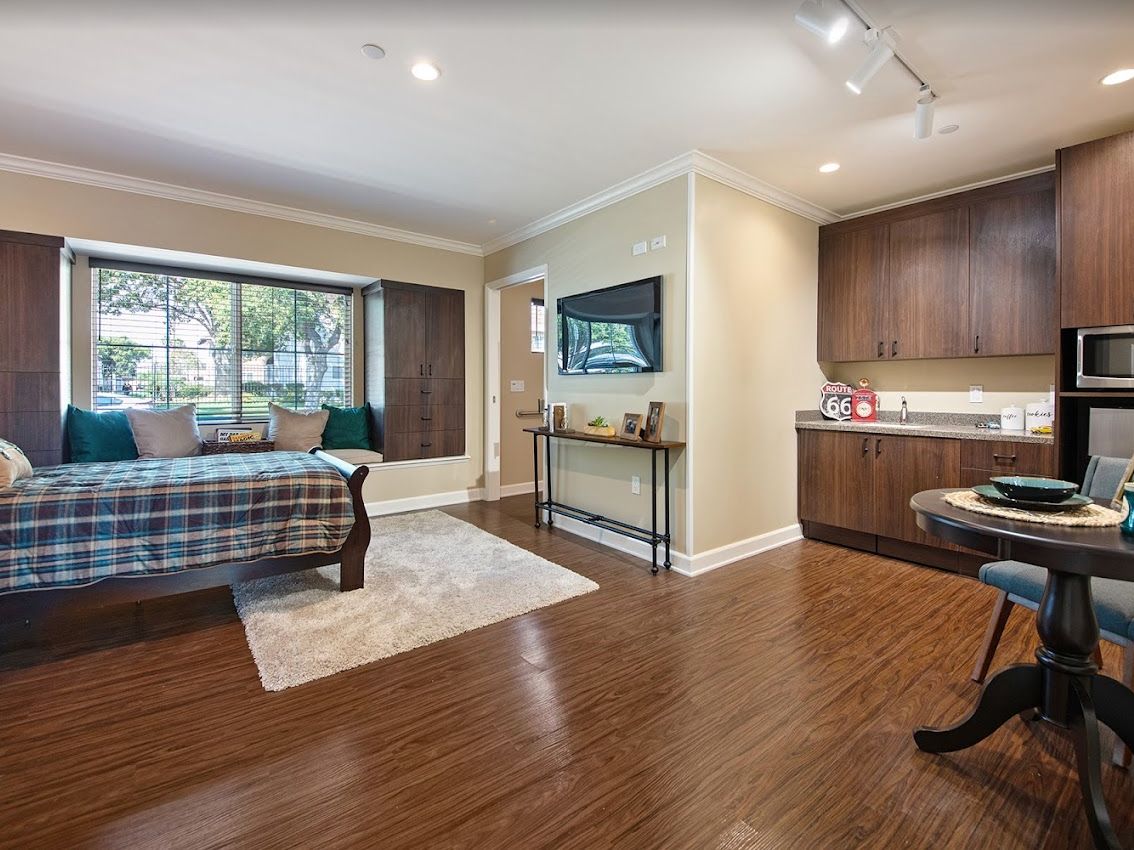 A modern open-space living area with hardwood floors, featuring a bed, a television on the wall, and a large window offering abundant natural light.