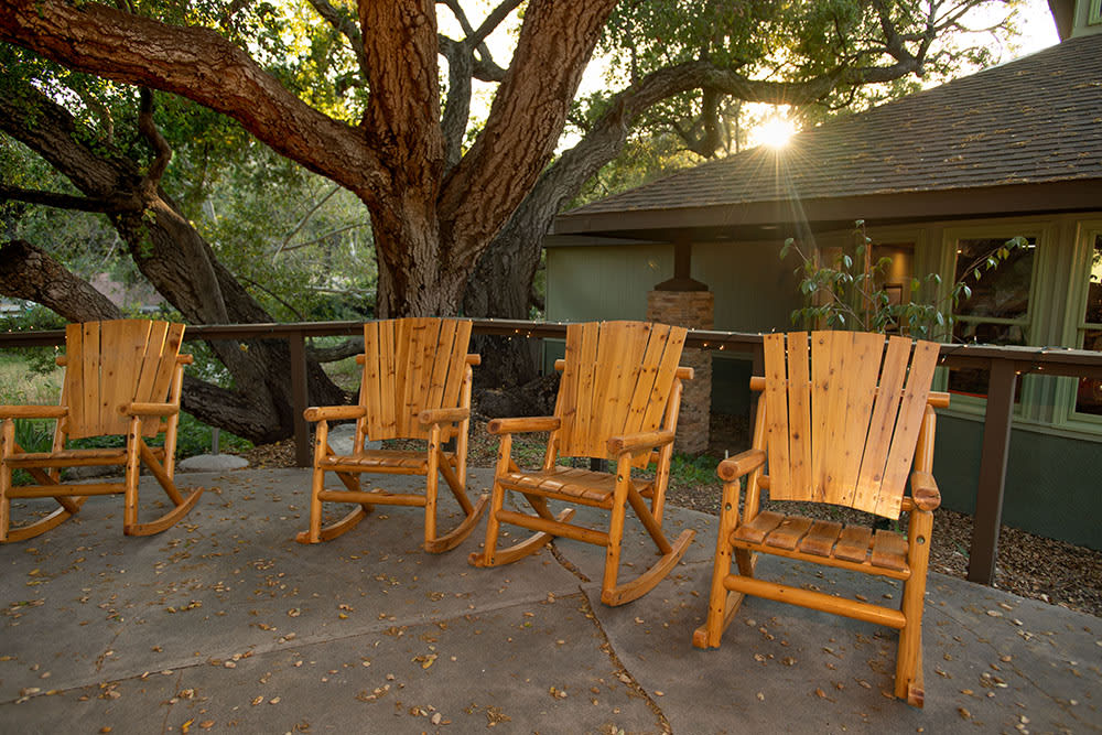 Wooden rocking chairs on a sunlit patio surrounded by trees.
