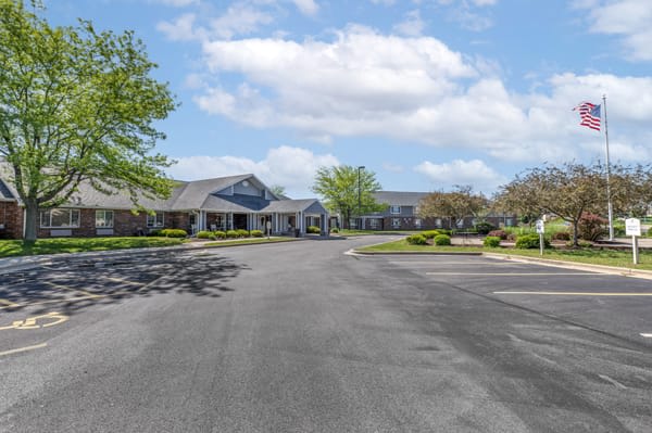 A view of a large building with a spacious driveway surrounded by trees under a partly cloudy sky.