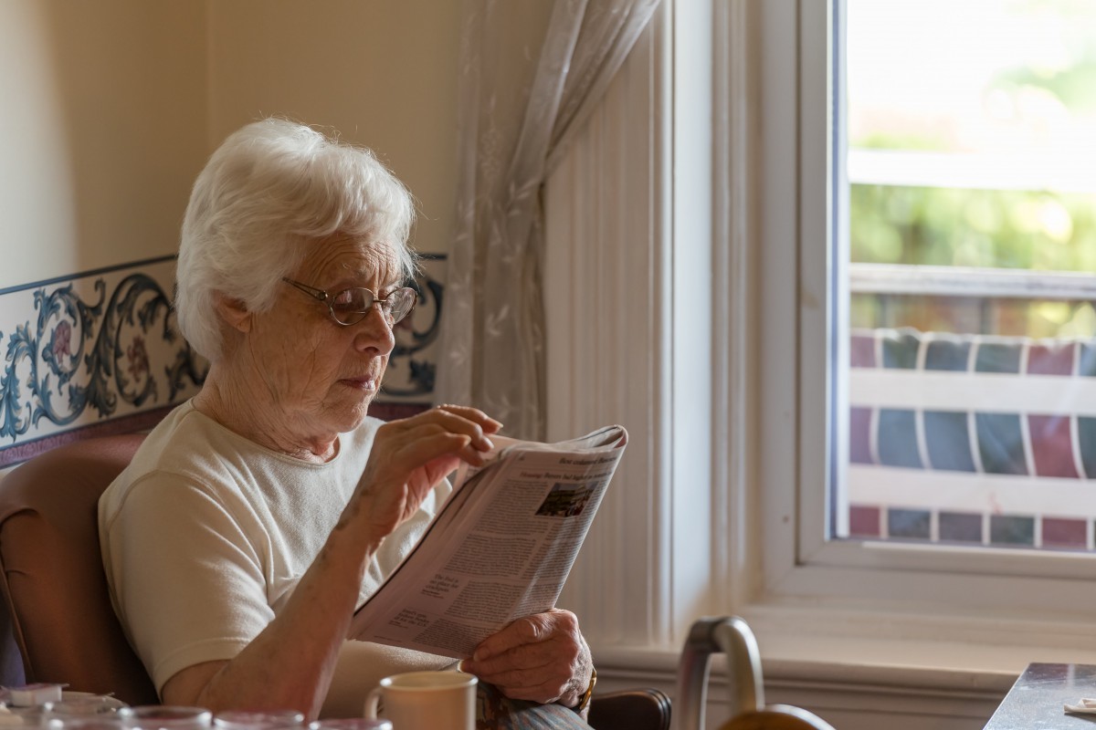 An elderly woman reads a newspaper by the window in daylight.