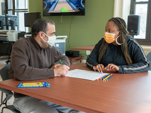 Two individuals engage in a discussion while working on a project at a table, both wearing masks.