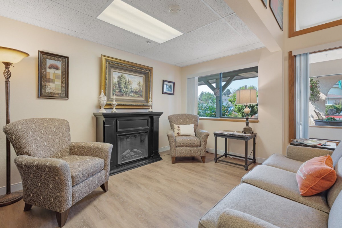 A cozy living room with comfortable armchairs and a fireplace, decorated with artwork and lit by a lamp near the window.