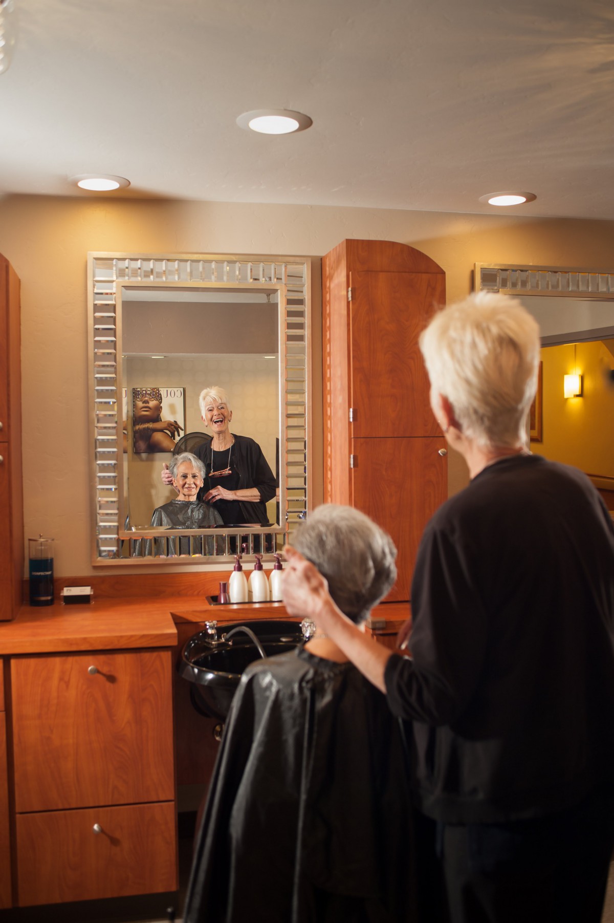 A hairdresser works on a senior woman's hairstyle in front of a mirror at a salon.