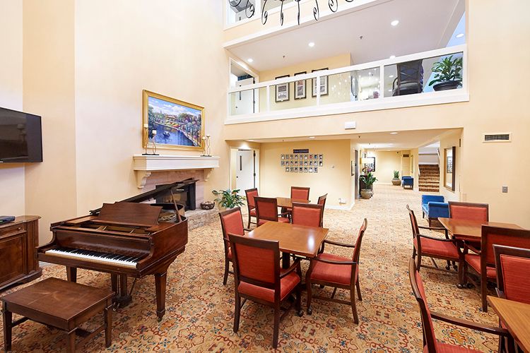 A spacious lobby area with a grand piano, tables, and chairs, decorated with artwork and a balcony overlooking the space.