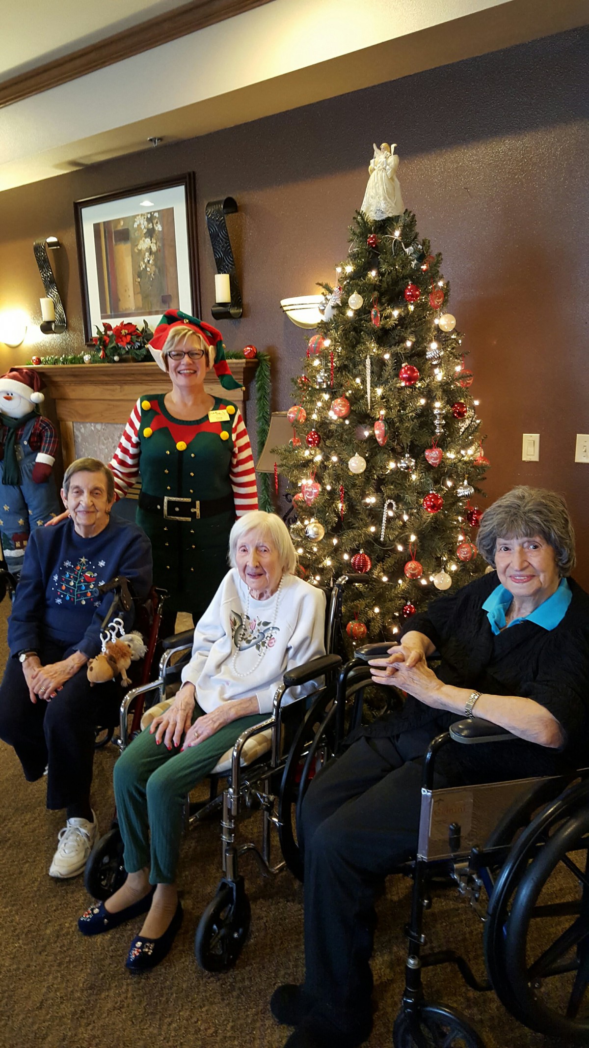 A group of people, including elderly individuals, gather around a decorated Christmas tree celebrating the holiday season.
