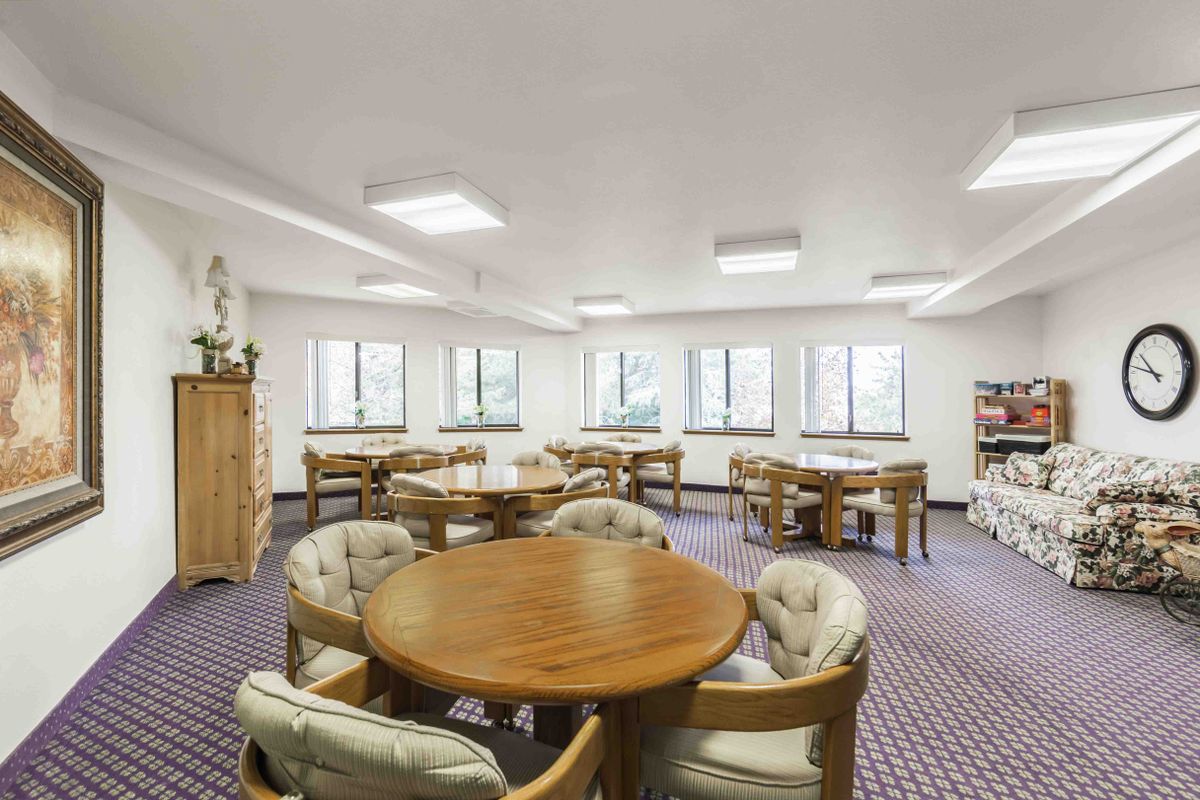 A spacious room with multiple round tables and cushioned chairs, featuring large windows and carpeted flooring.