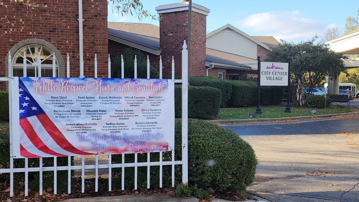 A fenced area with a building, featuring a banner and a sign surrounded by bushes.