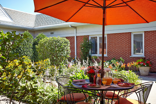 A charming outdoor dining space with a vibrant orange umbrella, surrounded by lush greenery and blooming flowers.