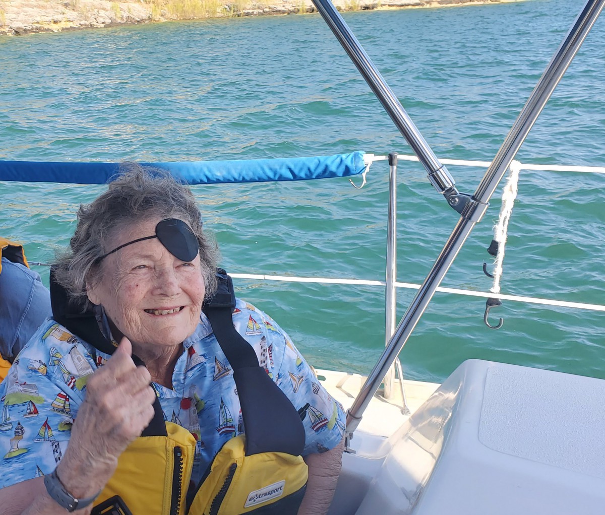 An elderly person wearing an eye patch and life jacket enjoys a sunny day on a boat.