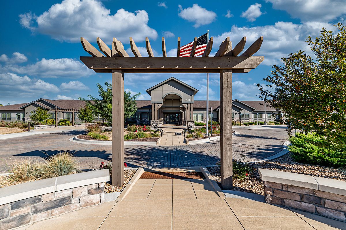 A pathway leads under a wooden pergola to a building with an American flag flying above.