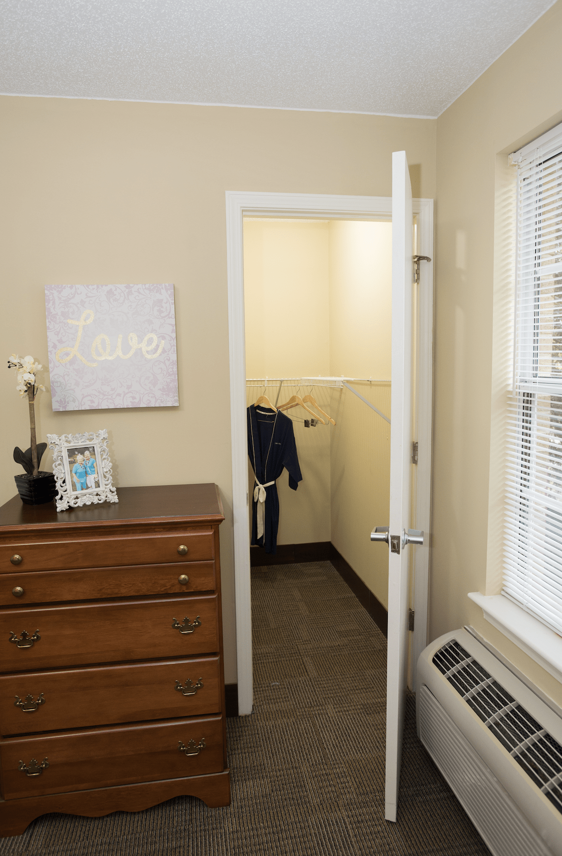 A cozy bedroom featuring a small closet with clothes hanging inside, a wooden dresser, and a window with blinds.