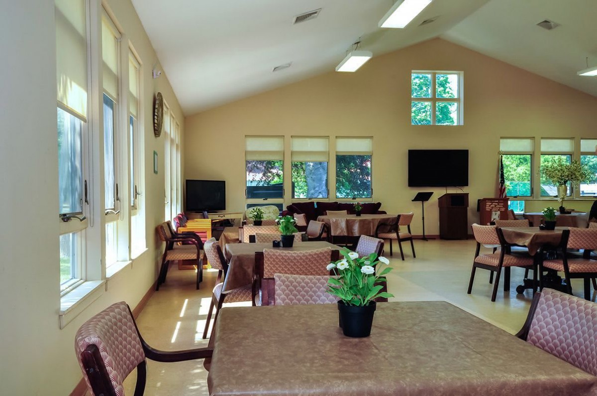A spacious community dining area with several tables, chairs, and potted plants, illuminated by large windows.