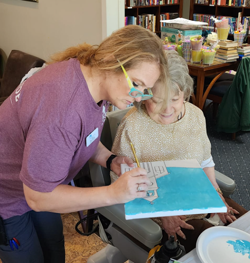 A caregiver assists an elderly person with a painting activity, promoting creativity and interaction.