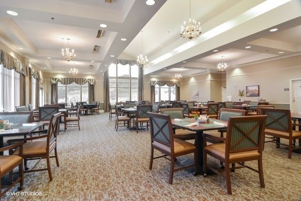 A spacious dining area with elegant chandeliers, featuring multiple tables and chairs on a patterned carpet.