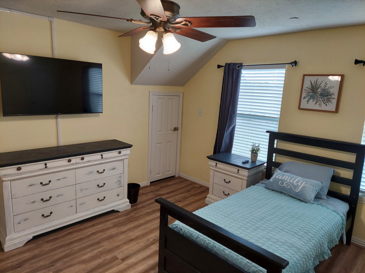 A cozy bedroom with a bed, dresser, wall-mounted TV, ceiling fan, and window with curtains.
