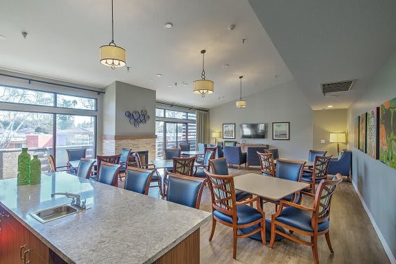 A cozy dining area with tables and chairs, featuring warm lighting and large windows.