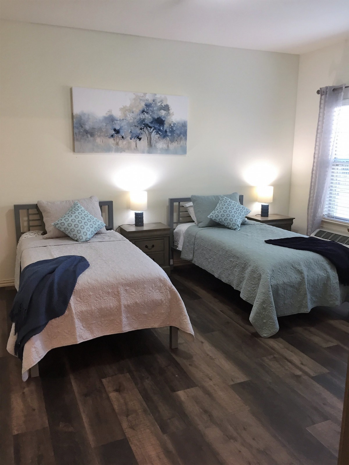 A cozy bedroom with two twin beds, decorated with pastel blankets and pillows, complemented by a large floral painting and warm bedside lamps.