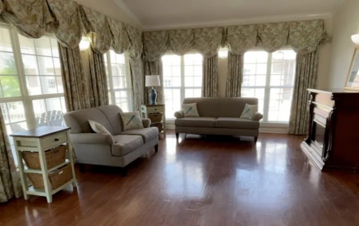 A spacious living room with elegant furniture, featuring a sofa and an armchair on a hardwood floor, adorned with large windows and decorative curtains.