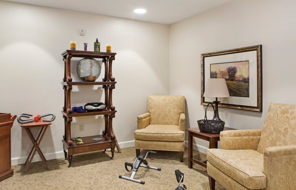 A cozy living room with comfortable chairs, a shelf holding decorative items, and small exercise equipment on the carpeted floor.