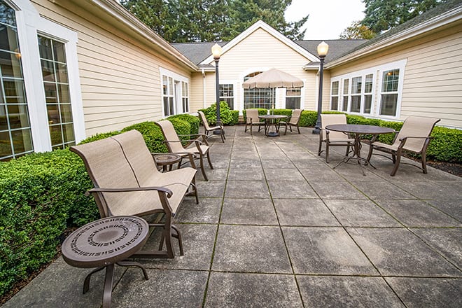 A spacious outdoor patio area with seating and tables, surrounded by a beige building and greenery.