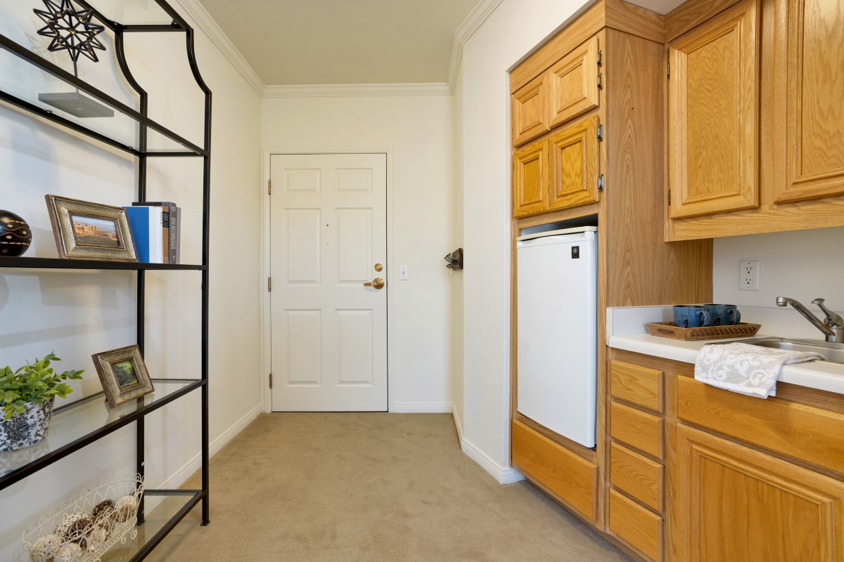 A kitchen area with wooden cabinets, a shelving unit, and a doorway in the background.