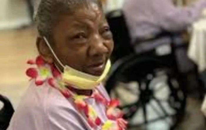 An elderly woman wearing a face mask and flower lei in a care setting.