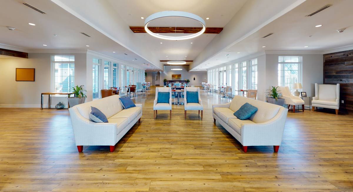 A spacious modern lounge with minimalist decor, featuring white sofas and blue cushions on wooden flooring, accentuated by large windows bringing in natural light.