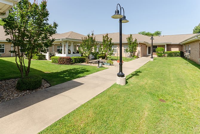 A well-manicured landscape with a paved pathway leading to a building, surrounded by green lawns and trees under a clear sky.