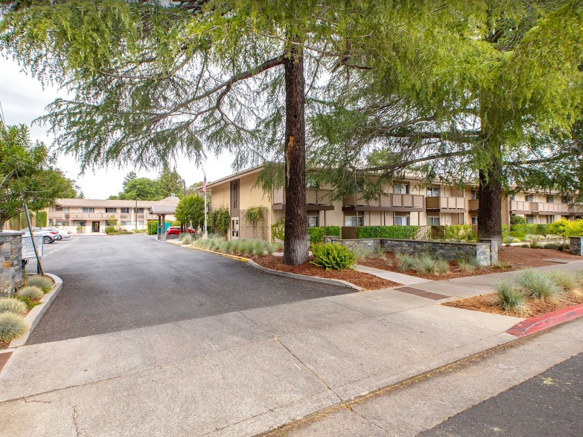 A residential complex surrounded by trees and greenery, featuring a driveway and sidewalk.