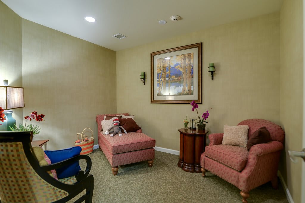 A cozy living room with a chaise lounge and matching armchairs, adorned with decorative pillows. A framed artwork hangs on the wall above, flanked by sconces. A small table holds a vase with flowers.