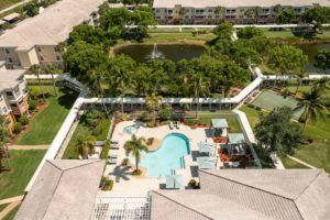 Aerial view of a residential area featuring a swimming pool, tennis court, and lush landscaping with palm trees and a pond.
