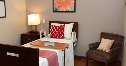 A neatly arranged hospital room featuring a patient bed with a red pillow, a bedside table, and a chair.