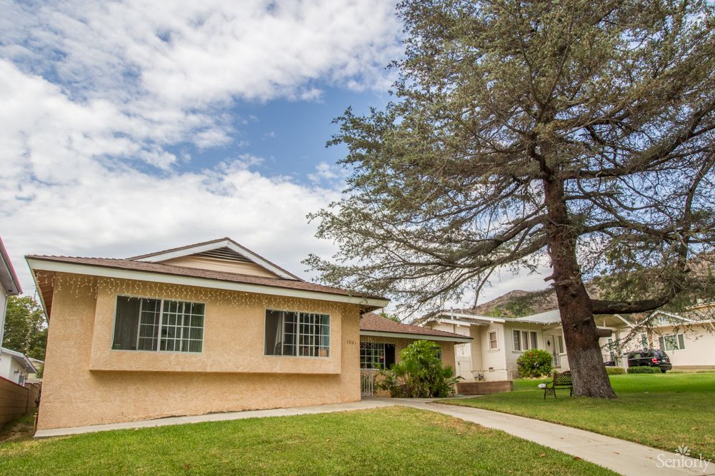 A suburban house with a large tree and a well-kept yard under a partly cloudy sky.