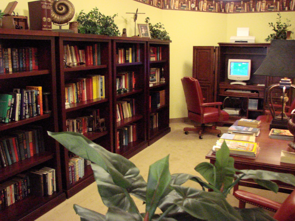 A cozy office space featuring bookshelves filled with books, a desk with a computer, and an armchair, all accented by indoor plants.
