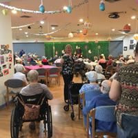 A community event at a nursing home with elderly residents seated, some in wheelchairs, participating in an indoor gathering with festive decorations.