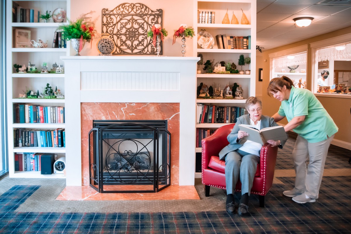 An elderly person reads with the assistance of a caregiver in a cozy living room setting featuring a fireplace and bookshelves.