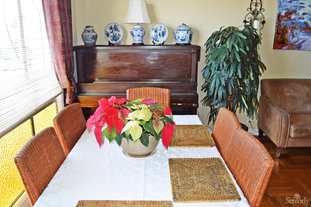 A dining room with a table set for four, featuring a poinsettia centerpiece and a piano in the background adorned with decorative vases.
