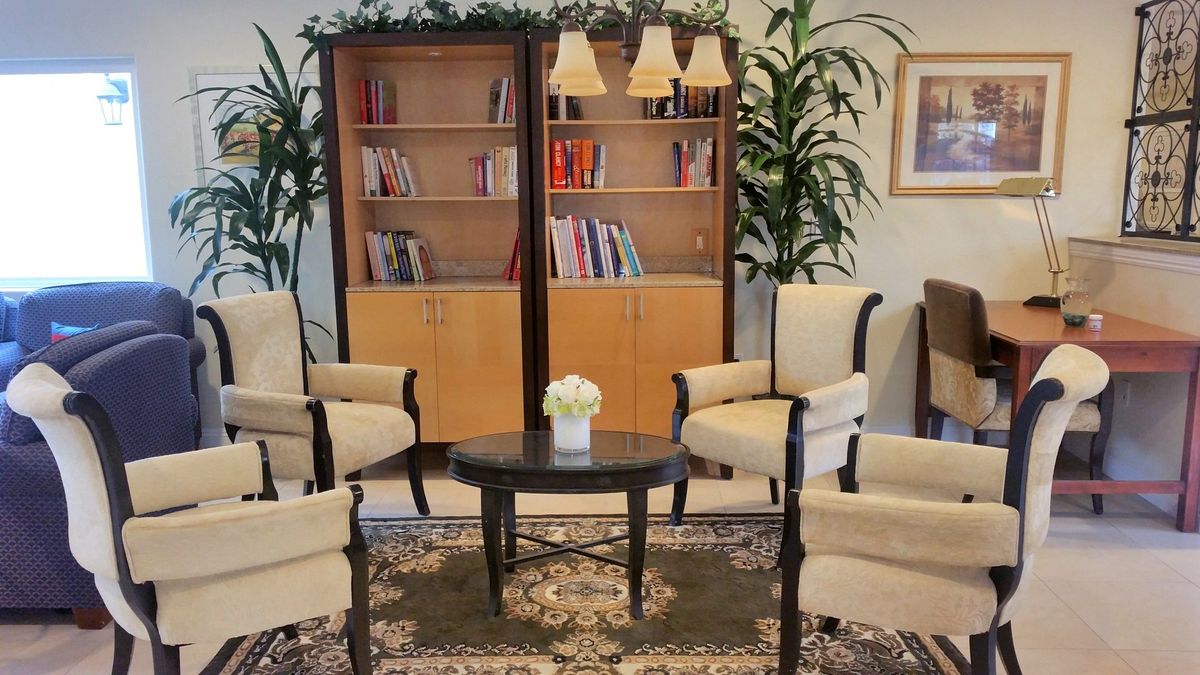 A cozy lounge area featuring armchairs, a central round table with flowers, bookshelves in the background, and indoor plants for decoration.