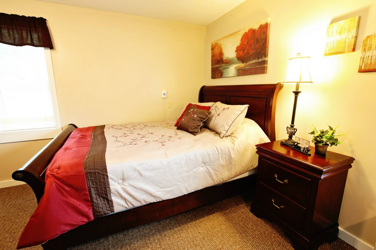 A cozy bedroom with a neatly made bed, warm lighting from a lamp on the nightstand, and a landscape painting on the wall.