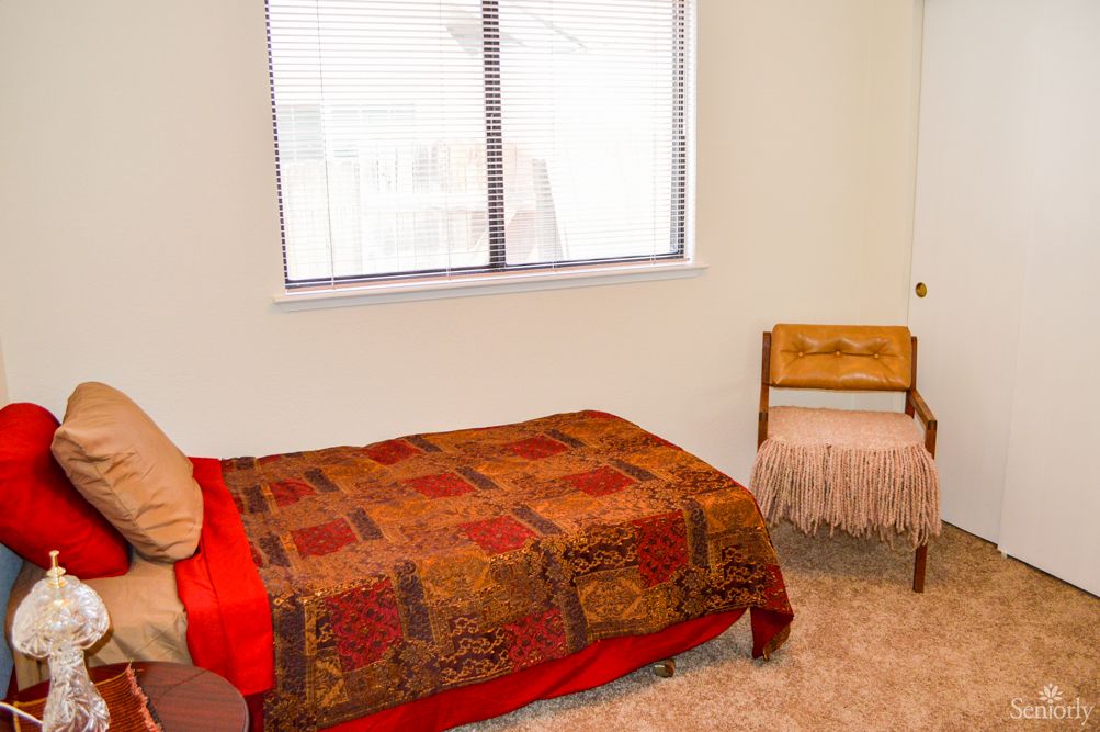A simple bedroom with a single bed, patterned quilt, and a wooden chair near the window.