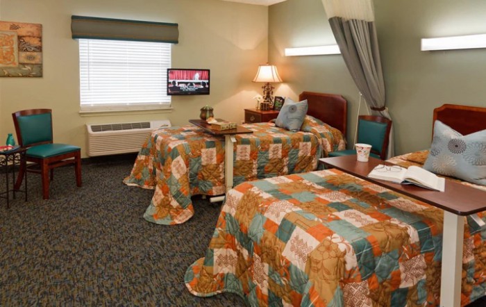 A hospital room featuring two beds adorned with colorful quilted bedspreads, a TV on the wall, and a window with blinds.
