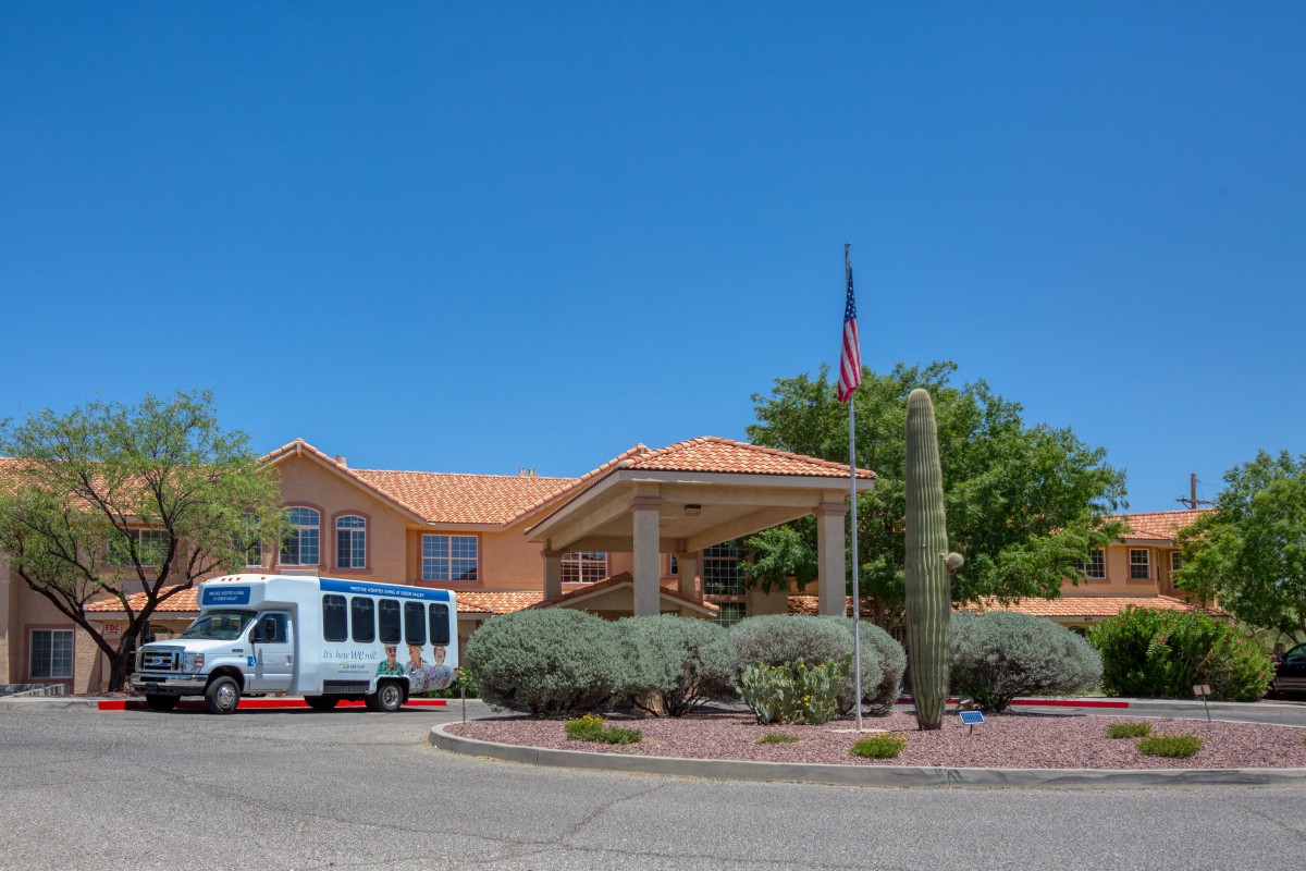 A hotel entrance with a shuttle bus parked nearby, surrounded by desert landscaping and a flag flying above.