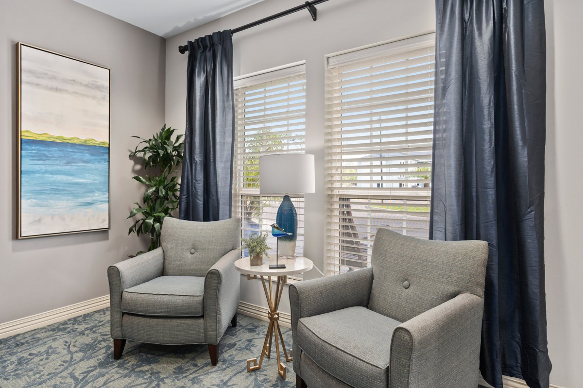 A cozy living room corner featuring two armchairs, a small table with a lamp, and large windows with blue curtains.